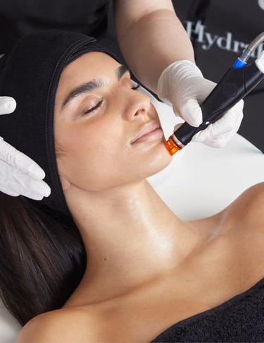 Woman receiving a facial Hydrafacial treatment with a device, wearing a black headband.
