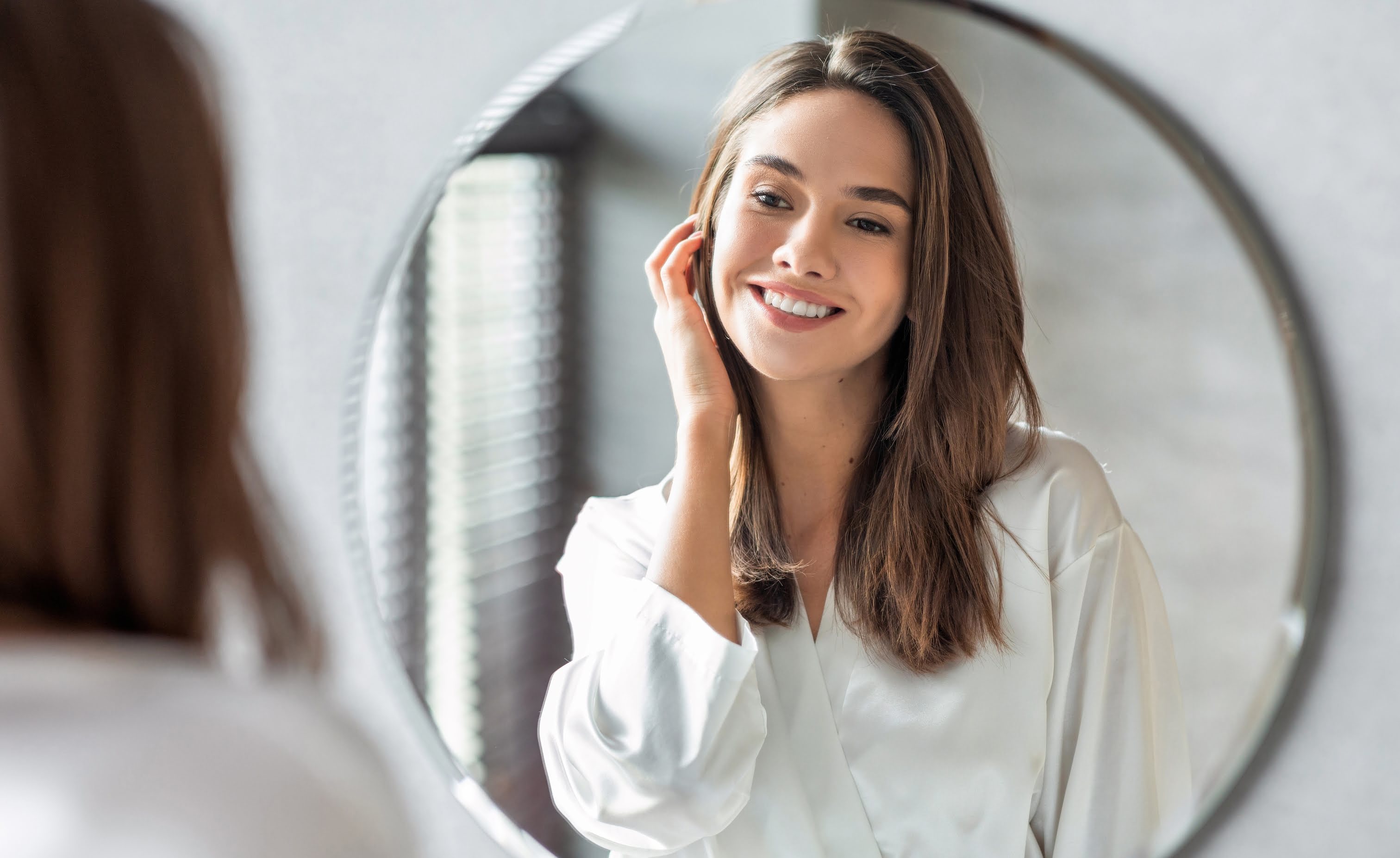 Woman looking at herself in the mirror, showcasing healthy skin, stronger nails, and shiny hair