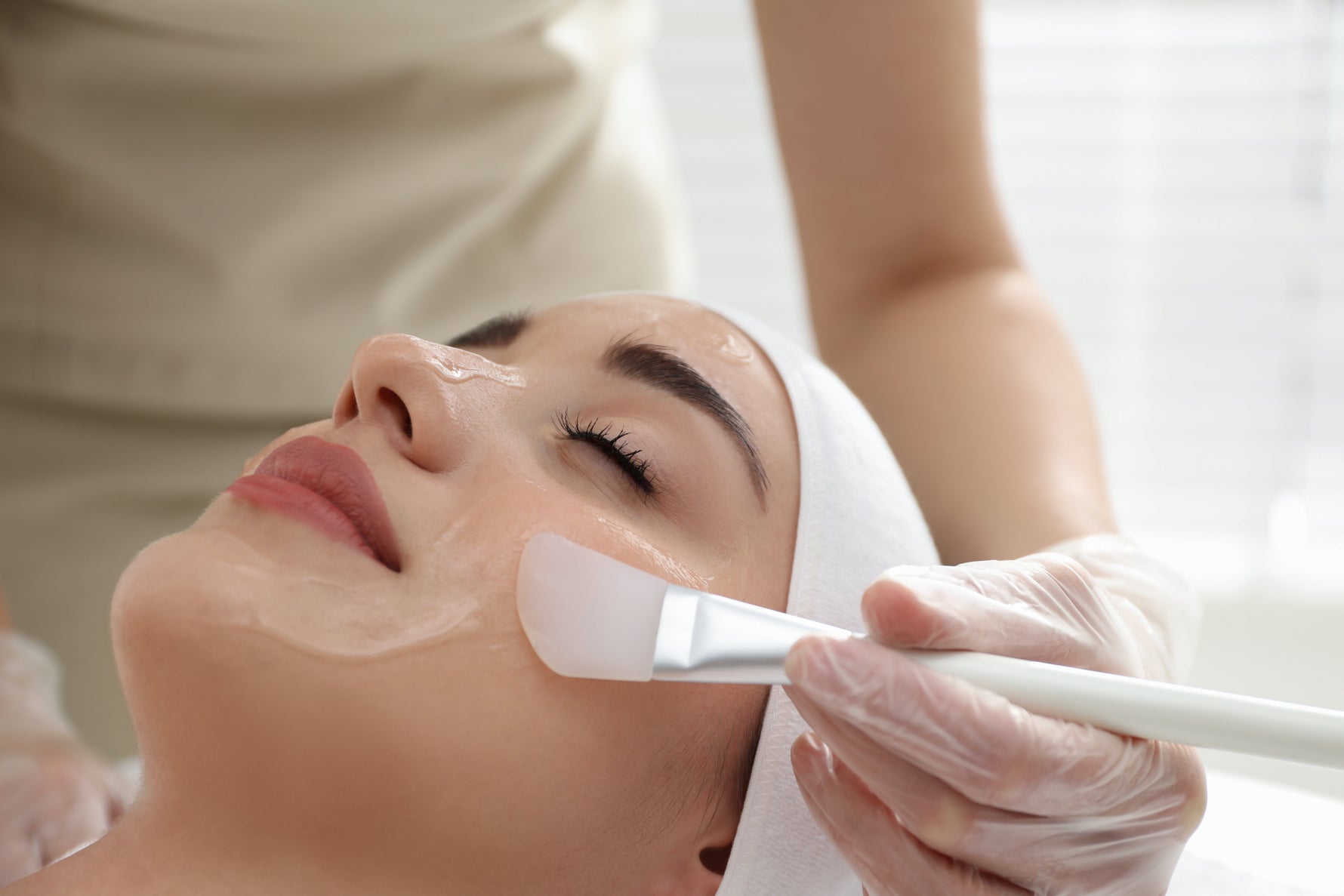 Aesthetic close-up of a chemical peel treatment being applied with a white translucent brush to a woman's face in a clean clinical setting